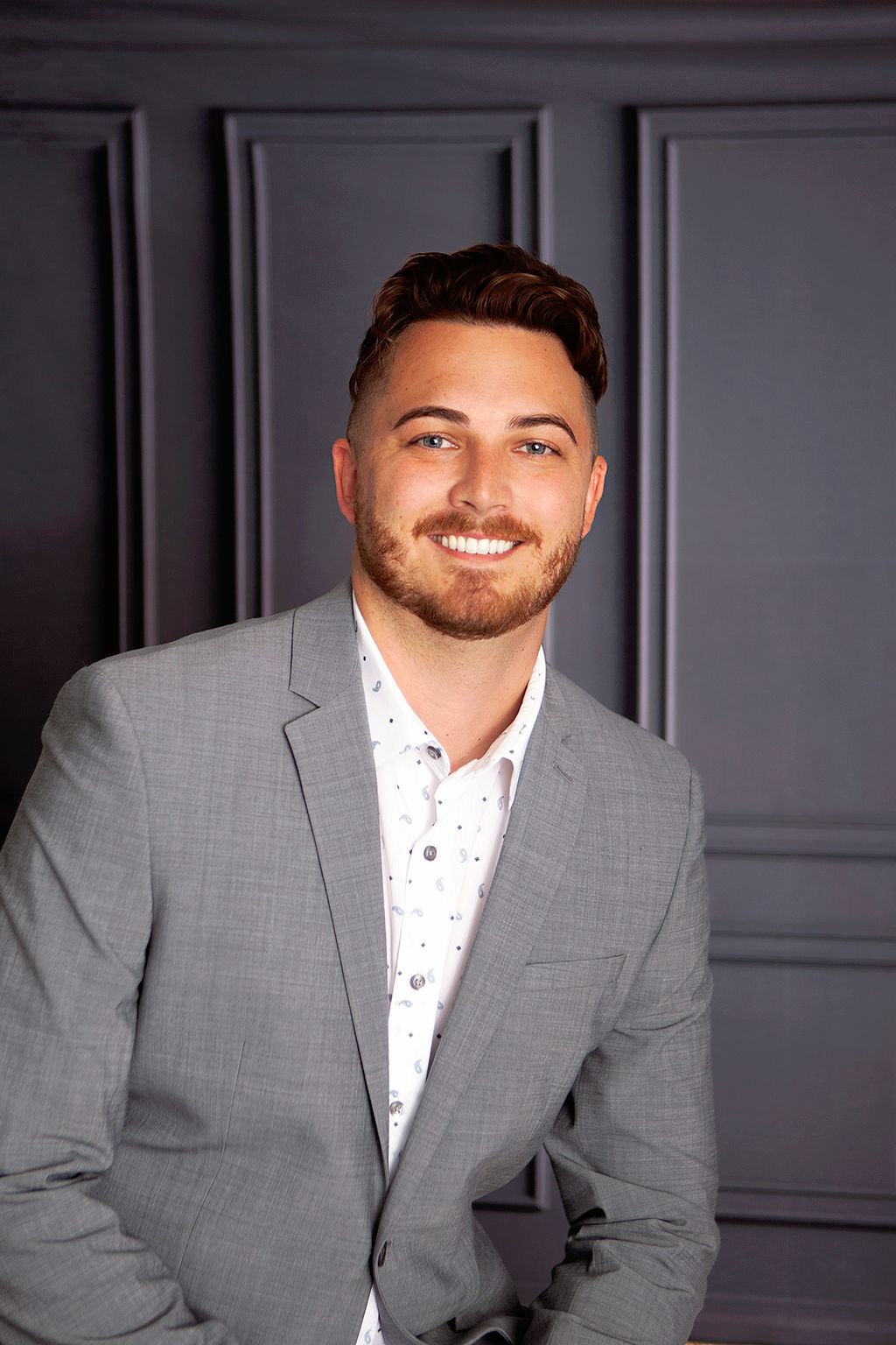 Professional headshot of a man in a gray suit against a dark paneled background